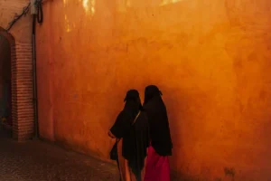 Two women in black walk down a sunlit alley.